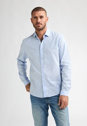 Mann mit kurzen Haaren und Bart, trägt ein hellblaues Hemd mit Knöpfen und blaue Jeans, steht mit einer Hand in der Tasche vor weißem Hintergrund.