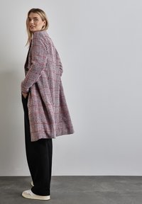 Lange geruite jas in rood en roomkleurige tinten, met een losse pasvorm, enkele knoopsluiting en een geweven stof met textuur. Combinatie met zwarte broek.