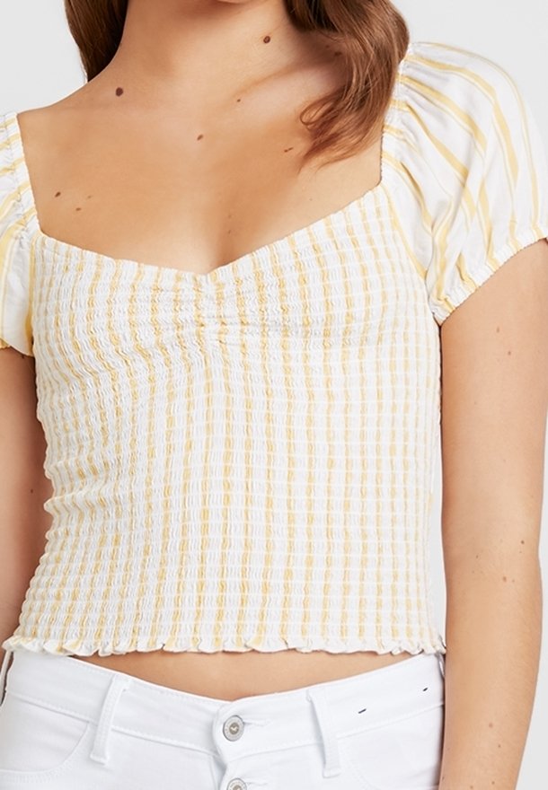 Biały top w paski w kolorze żółtym, krótki, z kwadratowym dekoltem, marszczonym materiałem, bufkami na rękawach i falbankowym wykończeniem. W połączeniu z białymi spodniami.