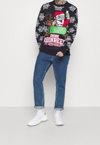 Pull en maille noire avec motif flocon de neige blanc, présentant un graphique de Père Noël et de renne avec un texte rouge et vert. Associé à un jean bleu retroussé et des baskets blanches.