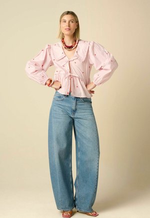 Femme portant un chemisier rose à manches bouffantes avec des détails floraux rouges, un jean bleu à jambes larges, des colliers superposés et des talons ouverts, debout les mains sur les hanches.