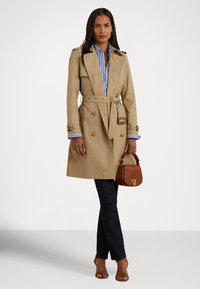 Μπεζ παλτό τραντς με διπλή σειρά κουμπιών, ζώνη στη μέση και επιθεώρητες στους ώμους. Συνδυασμένο με ριγέ πουκάμισο, σκούρα τζιν και καφέ τσάντα.