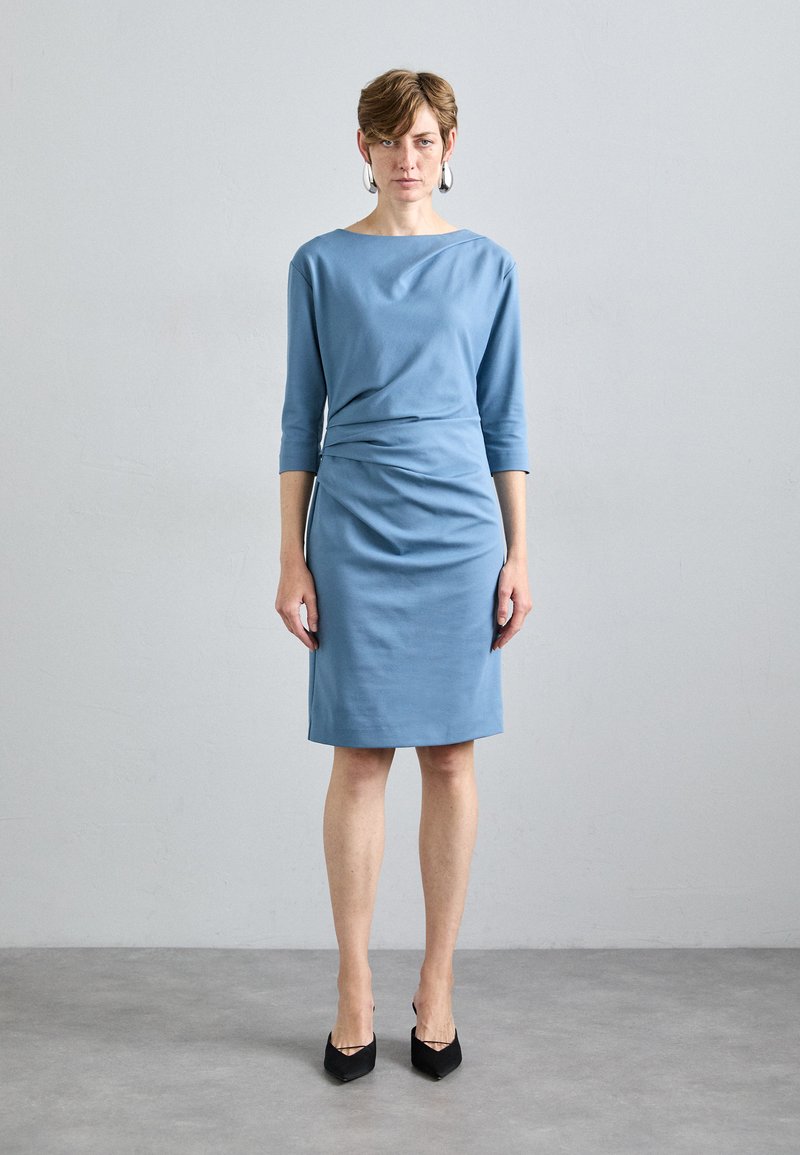 Vestido azul ajustado con mangas tres cuartos, detalle drapeado en la cintura y dobladillo a la altura de la rodilla. Llevado con tacones negros de punta.