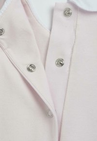 Vêtement rose pâle avec des boutons-pression argentés et un col blanc. Le tissu a une texture douce avec une finition lisse.