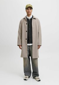 Beige uldoverfrakke med sorte knapper, lagd over en mørk sweater og hvid skjorte. Cargobukser i lys denim og cremefarvede sneakers fuldender looket.