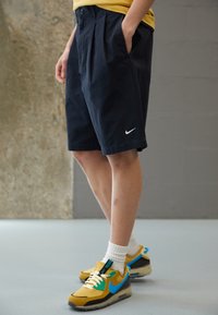 Black shorts with side pockets, pleated front, and white Nike logo. Worn with yellow and blue Nike sneakers and white socks.
