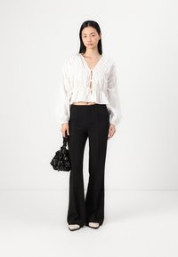 Blusa bianca corta con dettagli arricciati, scollatura profonda e maniche fluide abbinata a pantaloni neri a vita alta e una borsa nera con motivo.