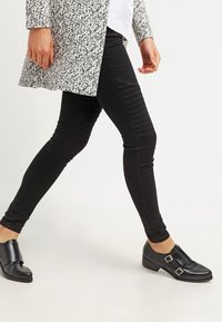 Jeans negros ajustados con detalles acanalados en los muslos, combinados con un abrigo gris estampado y zapatos negros con hebilla. Textura suave, ajuste a medida.
