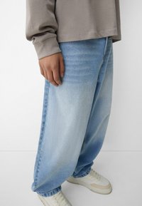 Hellblaue, locker geschnittene Jeans mit einem verblassten Muster, kombiniert mit weißen Sneakers und einem taupe farbenen Langarmshirt mit umgeschlagenen Ärmeln.