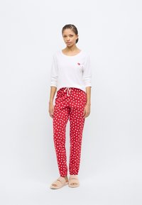 Λευκή μπλούζα μακρυμάνικη με μικρή κόκκινη καρδιά, συνδυασμένη με κόκκινο παντελόνι με λευκά σχέδια καρδιών και ελαστικές μανσέτες, φορεμένη με μπεζ χνουδωτές παντόφλες.