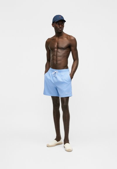 Atleetliku kehaga mees, kes kannab helesiniseid lühikesi pükse, valgeid libisevaid kingi ning tumesinist nokamütsi, seisab käed taskutes ühevärvilise tausta ees.