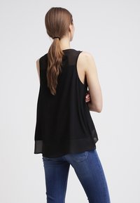 Blusa nera senza maniche con una parte superiore trasparente e un orlo inferiore solido. Il tessuto sembra leggero e leggermente testurizzato.
