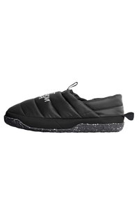 Musta pehmustettu liukuva kenkä, jossa on teksturoitu päällinen, speckled-mustalla pohjalla ja takana vetotappi. Logo-yksityiskohta päällisessä.