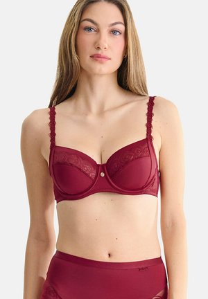 Femme aux longs cheveux blonds portant un soutien-gorge bordeaux à dentelle et une culotte taille haute assortie sur un fond blanc uni.