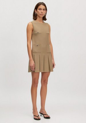Mouwloze beige jurk met twee voorzakken, zilveren knoopaccenten en een geplooide rok. Gemaakt van een gestructureerde stof.