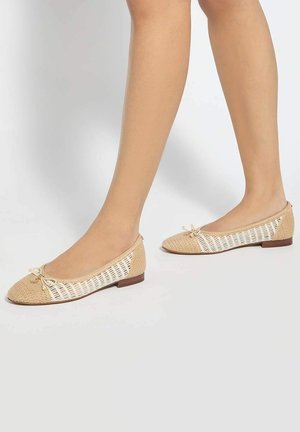 Legs wearing beige woven ballet flats with small bows, standing on a white surface against a plain background.