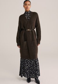 Smeđi pleteni kardigan s dubokim V izrezom, prednjim džepovima i crnim remenom s zlatnim detaljem. Nosit će se preko crne haljine s točkastim uzorkom.