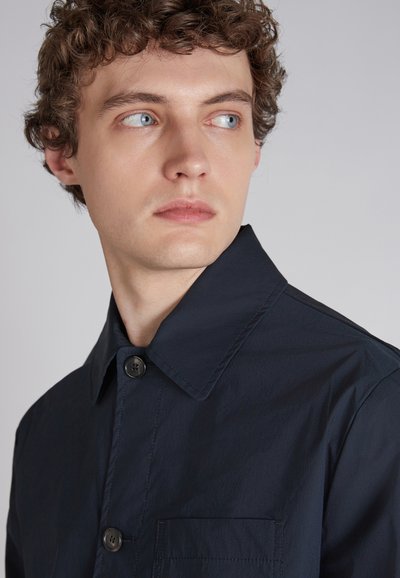 Granatowa koszula zapinana na guziki z klasycznym kołnierzykiem, kieszonką z przodu i strukturalną tkaniną. Dwa guziki widoczne na listwie.