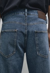 Dark blue denim jeans with a high waist, featuring two back pockets and contrasting stitching, paired with a brown leather waistband label.