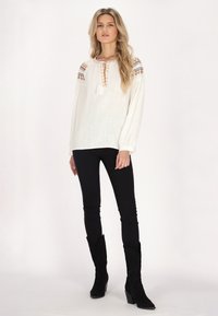 Bílá halenka s dlouhým rukávem a vyšívaným vzorem na ramenou, s výstřihem do V a zavazováním, vyrobená z texturované látky. Černé skinny džíny a boty.
