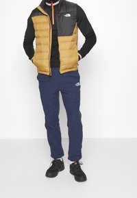 Gepolstertes schwarzes und goldenes Weste über einem schwarzen Langarmshirt, kombiniert mit navyblauen Jogginghosen und schwarzen Sneakers. Auffällige Markenlogos sind sichtbar.