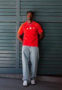 Rode sportieve shirt met oranje strepen en logo, gecombineerd met lichtblauwe jeans en witte sneakers. Staand tegen een groene lattenwand.