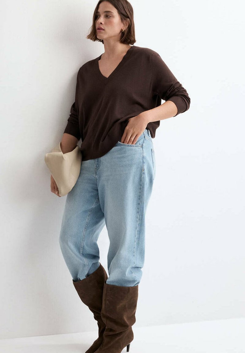 Femme aux cheveux courts portant un haut à manches longues marron foncé, un jean bleu clair et des bottes marron larges, tenant une pochette beige.