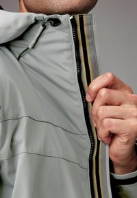 Jachetă gri cu un finisaj texturat, având un fermoar dublu și un accent de țesătură în dungi în interior. Mână apucând gulerul.