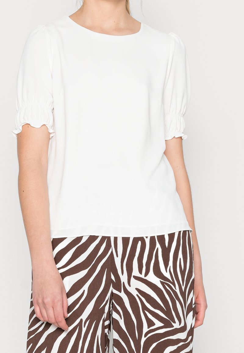 Blouse blanche avec manches courtes froncées, encolure ronde et texture lisse, assortie à un pantalon à rayures zébrées marron et blanc.