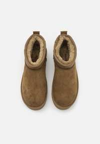 Botas de ante marrón hasta el tobillo con un exterior texturizado y un suave forro de piel sintética, que presentan un diseño sencillo y una tira en la parte posterior para facilitar el calzado.