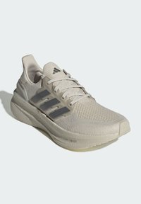 Zapatilla de running Adidas color beige claro con parte superior de punto, plantilla acolchada, cierre de cordones y tres rayas laterales gris oscuro.