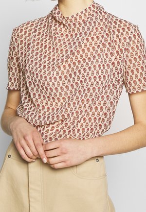 Kvinde iført en kortærmet, højhalset bluse med et rødt og beige paisley-mønster, stoppet ned i beige bukser.