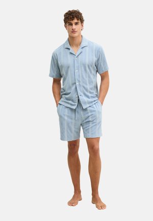 Man staat op blote voeten, draagt een lichtblauw overhemd met korte mouwen en bijpassende korte broek met verticale strepen, handen in de zakken.
