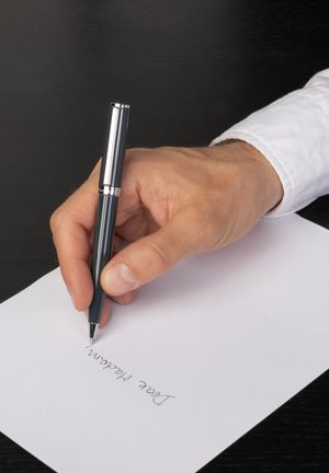 Mano destra che tiene una penna, scrivendo "Gentile Signora" su un foglio bianco contro una superficie nera.