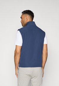 Mörkblå ärmlös väst med hög krage, tillverkad av ett mjukt material. Har platta sömmar och en avslappnad passform, bärs över en vit T-shirt.