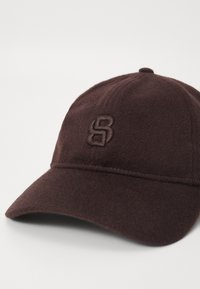 Donkerbruine wollen baseballcap met gebogen rand en geborduurd logo van op elkaar verweven letters op het voorpaneel, weergegeven op een witte achtergrond.