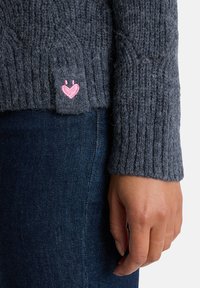 Grauer gestrickter Pullover mit einem gerippten Muster und einem rosa bestickten Herzdetail am Saum. Die Hand des Models ist sichtbar, das blaue Jeans trägt.