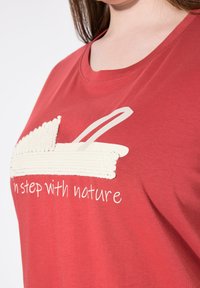 Camiseta de algodón roja con un diseño de cuerda blanca texturizada y la frase impresa "en sintonía con la naturaleza" debajo del detalle gráfico.