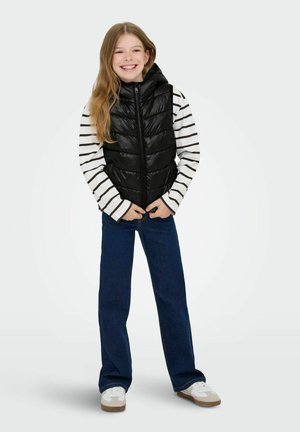 Gilet en doudoune noir avec capuche sur une chemise à rayures noir et blanc à manches longues, associé à un jean en denim foncé et des baskets blanches.