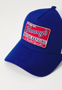 Blå basebollkeps med vit nätbak. Har ett broderat märke med "Tommy's Motor Reparations" och röda detaljer. Inscription "100% äkta delar".