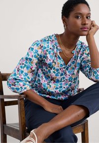 Blusa floreale in tonalità rosa, blu e verde; presenta un colletto a frange e maniche a tre quarti. Abbinata a pantaloni navy e scarpe chiare.