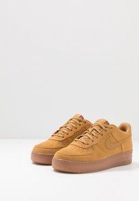 Baskets en suedine beige avec une zone perforée sur le bout, des lacets et le logo Nike sur le côté. Semelle en caoutchouc translucide. Design arrondi avec des accents subtils.
