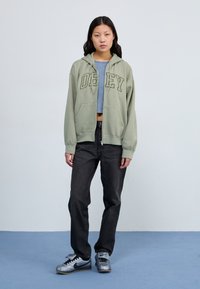 Green zip-up hoodie with "OBEY" text, paired with high-waisted black jeans and silver Nike sneakers. Smooth fabric with a relaxed fit.