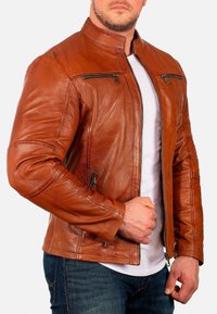 Bruine leren jas met een staande kraag, rits aan de voorkant en twee borstzakken. Getailleerd ontwerp met zichtbare stiksels op de schouders en mouwen.