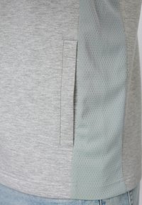 Γκρι ύφασμα με κατακόρυφη τσέπη με σχισμή δίπλα σε ανοιχτό πράσινο διαπνέον πλέγμα, φορεμένο πάνω από τζιν παντελόνι ανοιχτού γαλάζιου χρώματος.