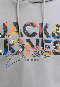 Sudadera gris con un atrevido estampado gráfico multicolor que incluye el texto "JACK & JONES Essentials" y el año de establecimiento 1990.