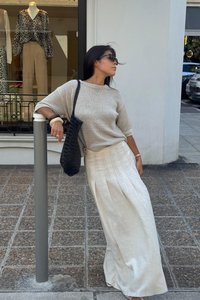 Maglione grigio chiaro a maglia, gonna plissettata crema, borsa intrecciata nera. Tessuti morbidi, vestibilità rilassata e stile casual sullo sfondo urbano.