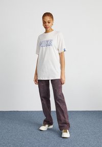 Krémové tričko Nike s modrým logem a volným střihem. Kombinováno s širokými kalhotami tmavě fialové barvy. Bílé tenisky s hnědými tkaničkami. Jednoduchý design.