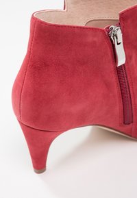 Bottine talon en daim rouge avec fermeture éclair latérale argentée, talon effilé bas, et doublure intérieure beige clair sur fond blanc.