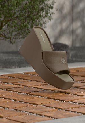Καφέ πλατφόρμα σανδάλι Calvin Klein με υφασμάτινη λουρίδα και ανάγλυφο λογότυπο «ck» σε ξύλινο δάπεδο σε εξωτερικό χώρο.